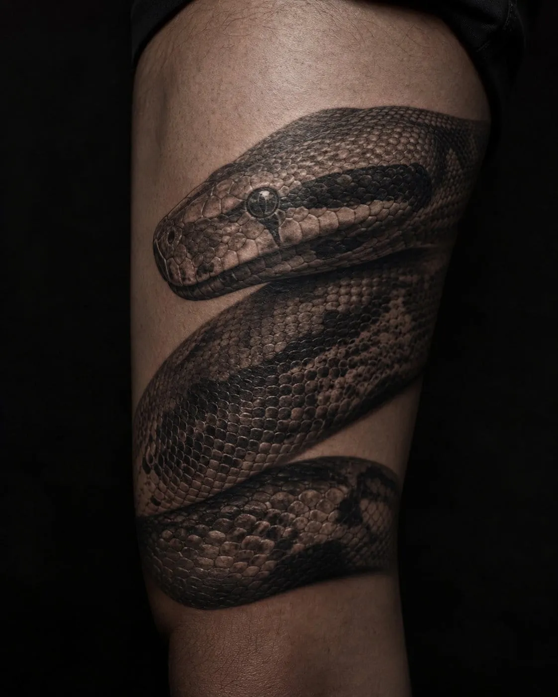 Photorealistic black-and-grey python tattoo with detailed scales and a glinting eye on a thigh.